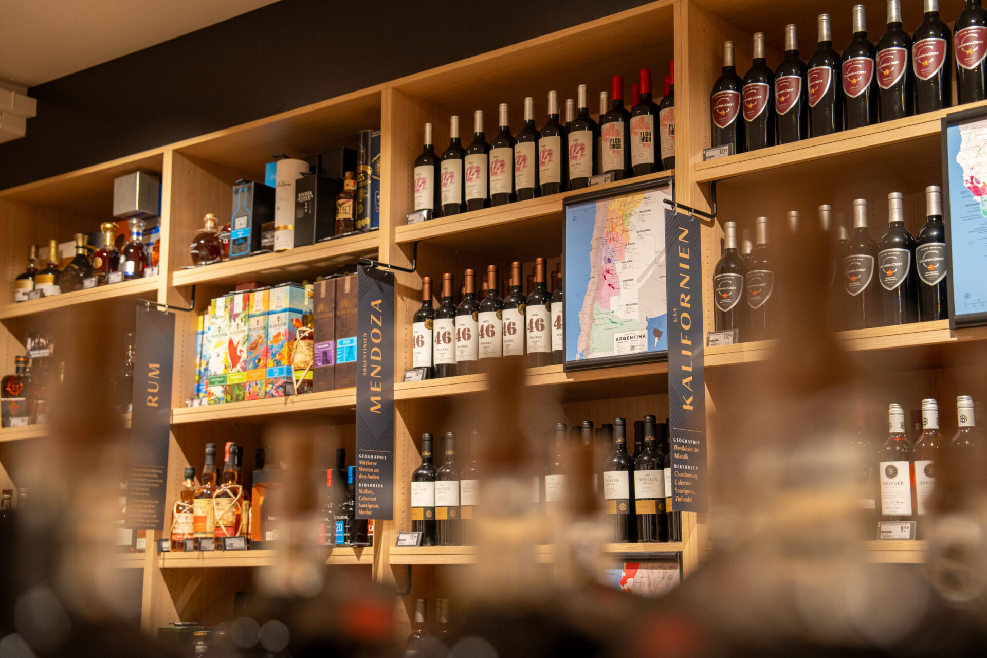 Regale mit verschiedenen Flaschen Wein und Spirituosen, beschriftet mit „Rum“, „Mendoza“ und „Kalifornien“.