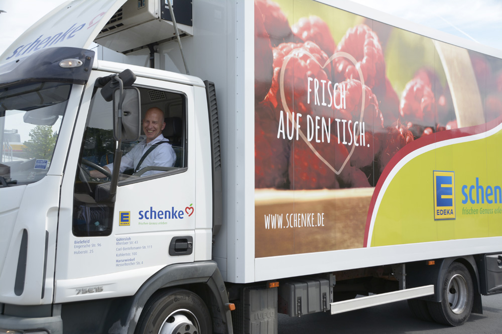 Ein LKW von Schenke mit der Aufschrift "Frisch auf den Tisch" und Werbung für EDEKA fährt auf der Straße.