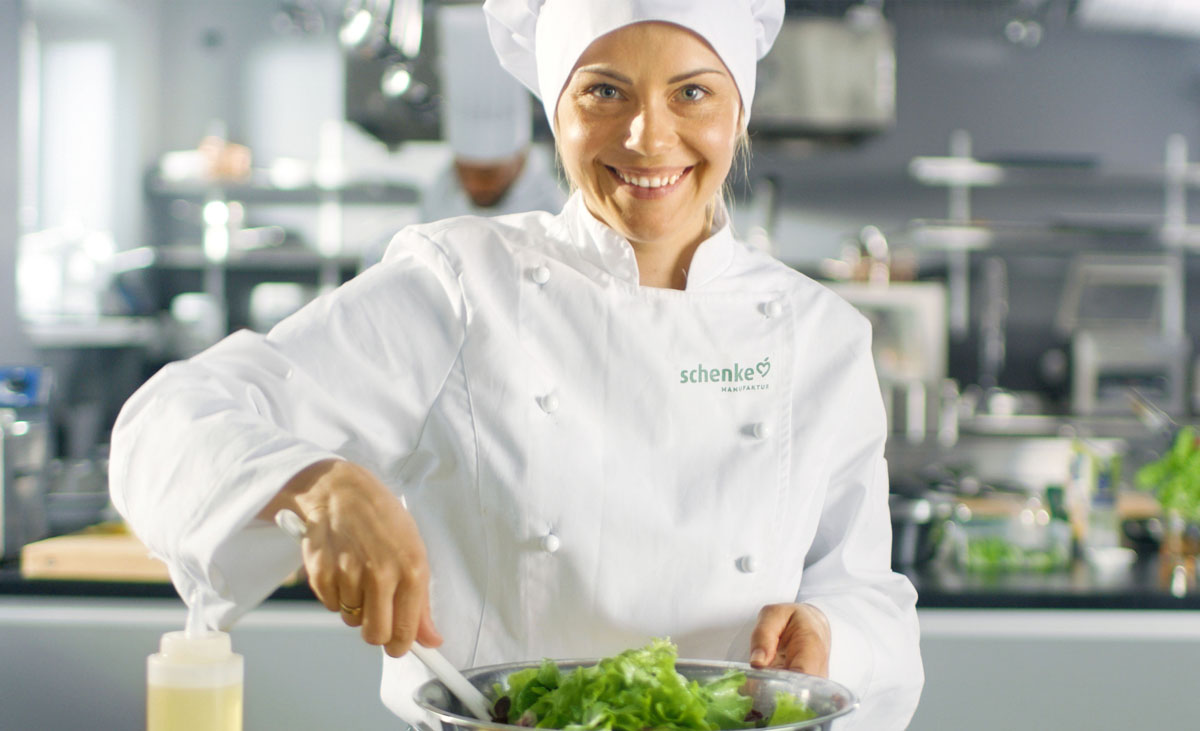 Köchin in weißer Uniform, die Salat in einer Schüssel zubereitet und lächelt.