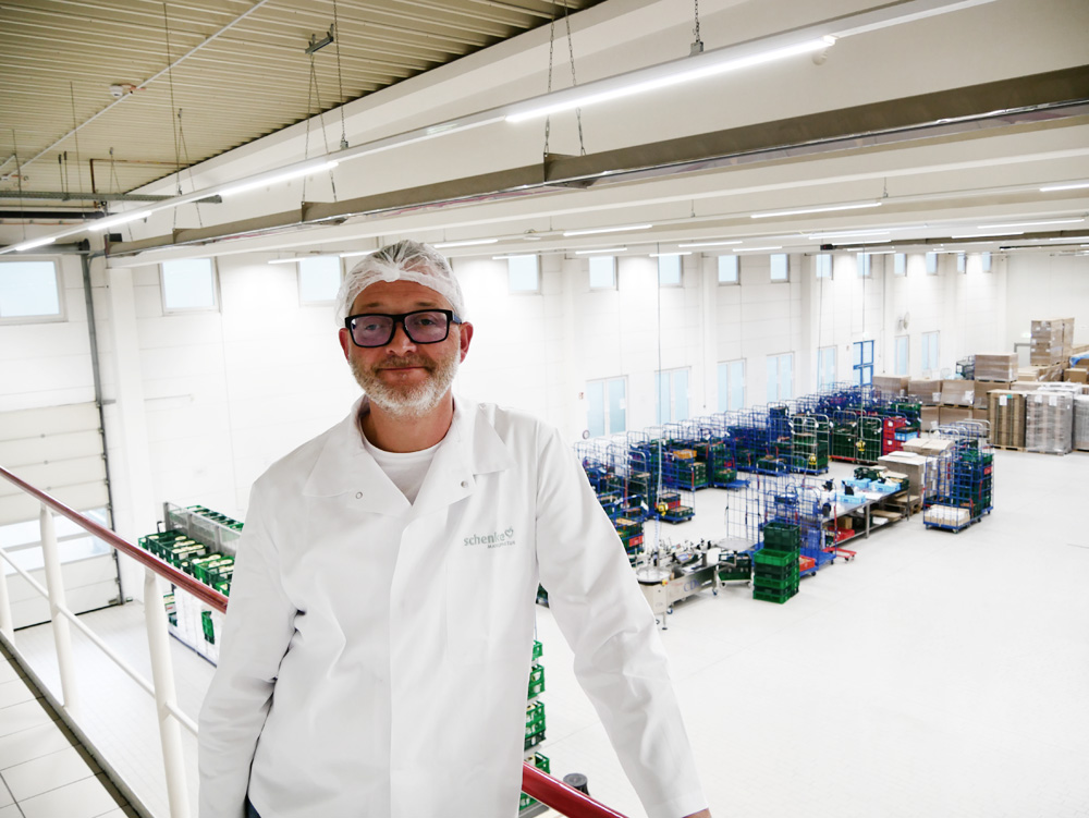 Person in weißen Kittel und Schutzhaube in einer Fabrikhalle mit Regalen und Kisten im Hintergrund.