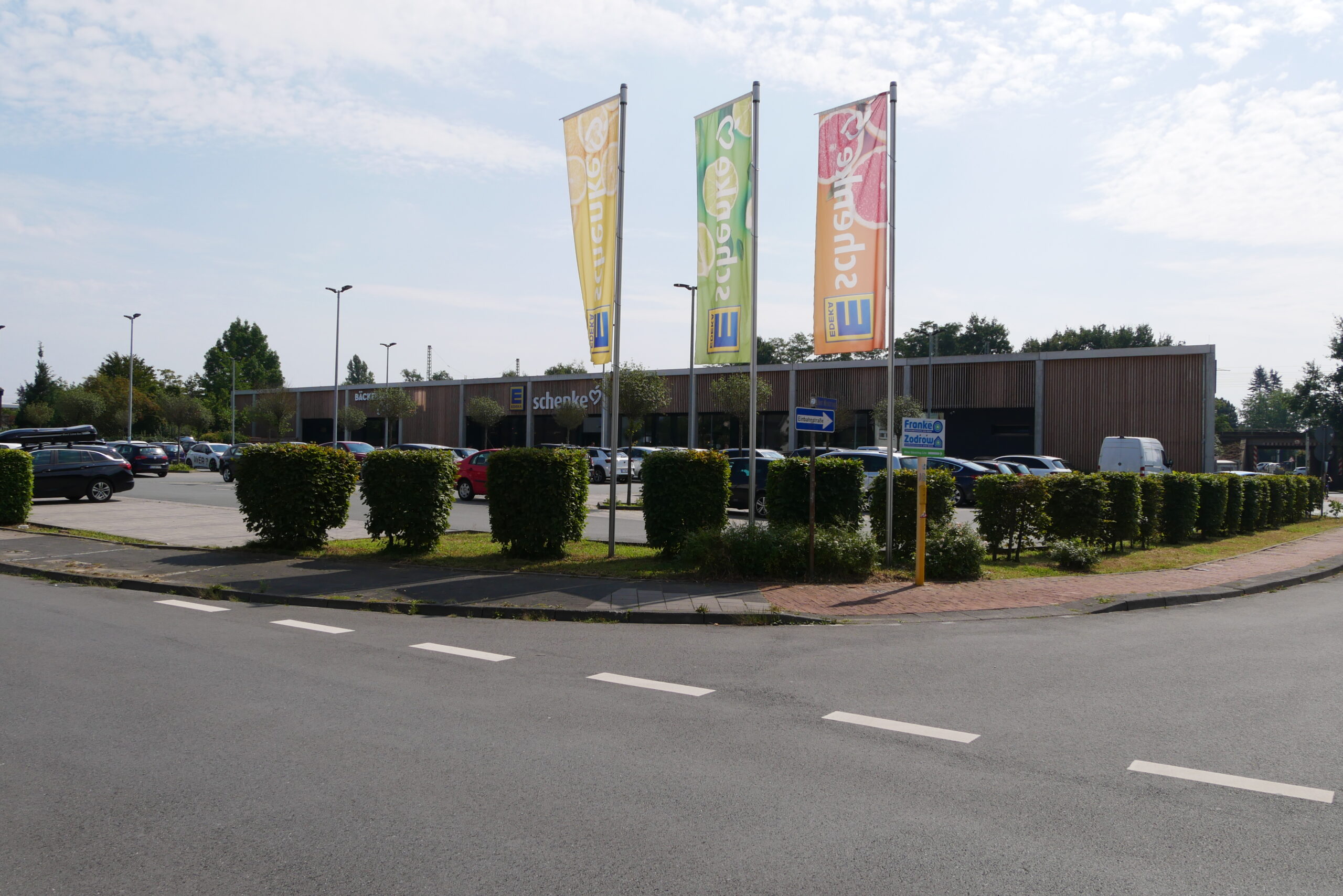 Einkaufszentrum mit mehreren Flaggen, geparkten Autos und grüner Umgebung.