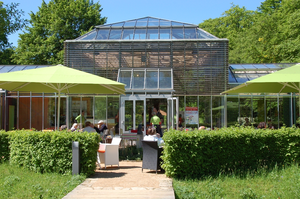 Ein modernes Gewächshaus mit Café, umgeben von grünen Pflanzen und Sonnenschirmen, Menschen sitzen im Freien.