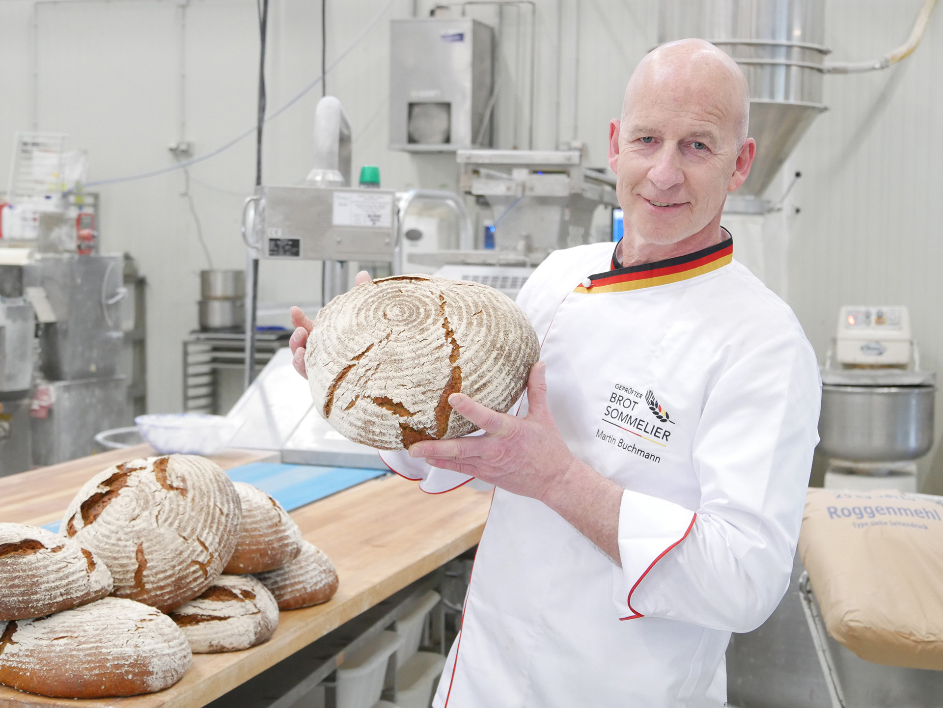 Ein Mann in weißer Bäckerjacke hält ein rundes handwerklich hergestelltes Brot in einer Bäckerei mit mehreren Broten auf einem Holztisch im Hintergrund.