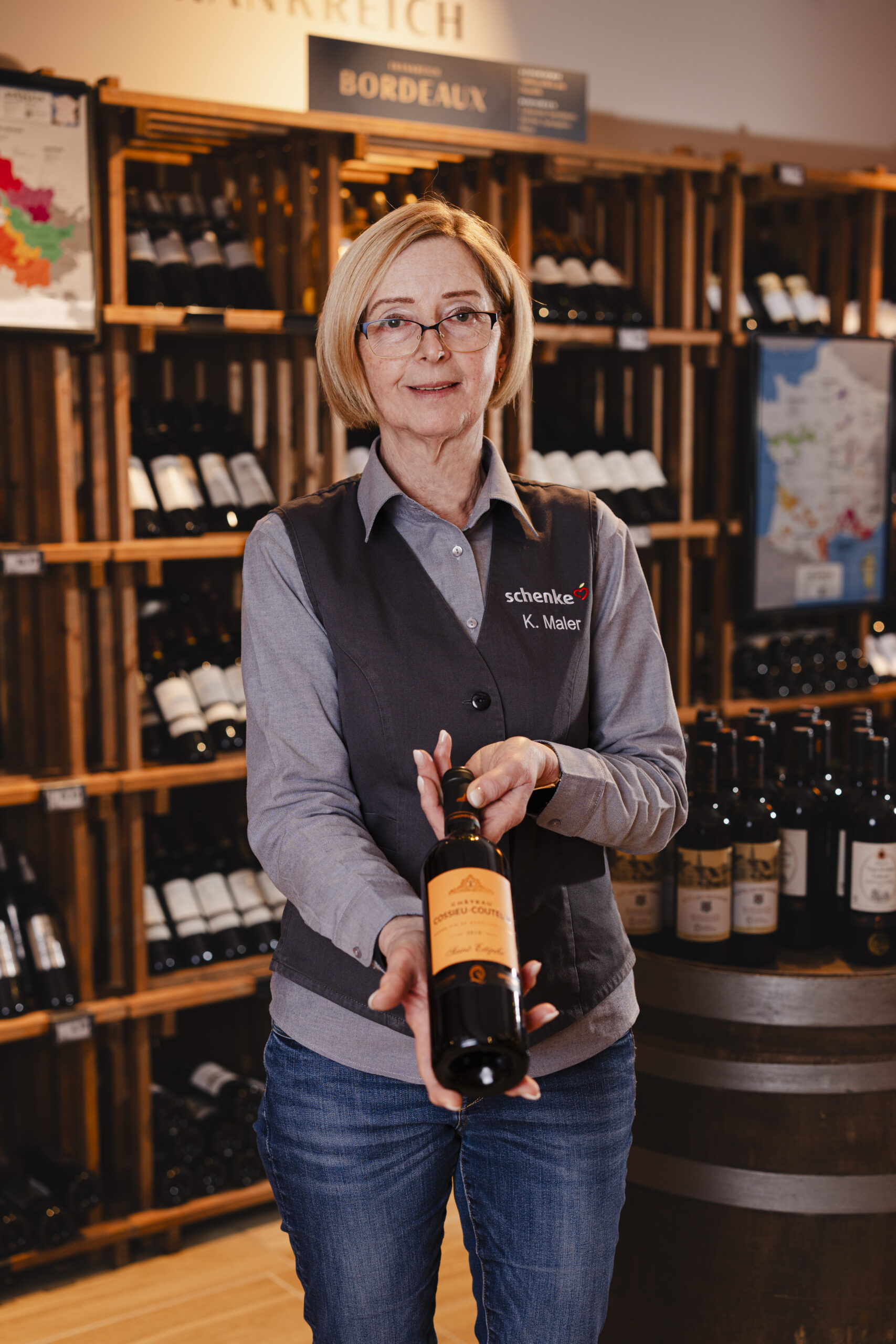 Eine Frau in einer Weinhandlung hält eine Flasche Wein in den Händen, vor einem Regal voller Weinflaschen. Im Hintergrund ist ein Schild mit der Aufschrift "Bordeaux" sichtbar.