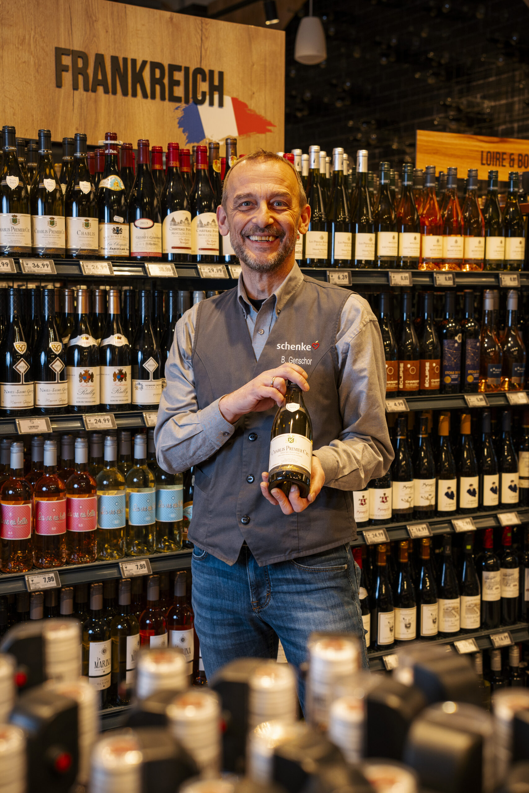 Ein Mitarbeiter mit grauer Weste hält eine Weinflasche in einem Supermarkt vor einem Regal mit französischen Weinen, beschriftet mit 'Frankreich'.
