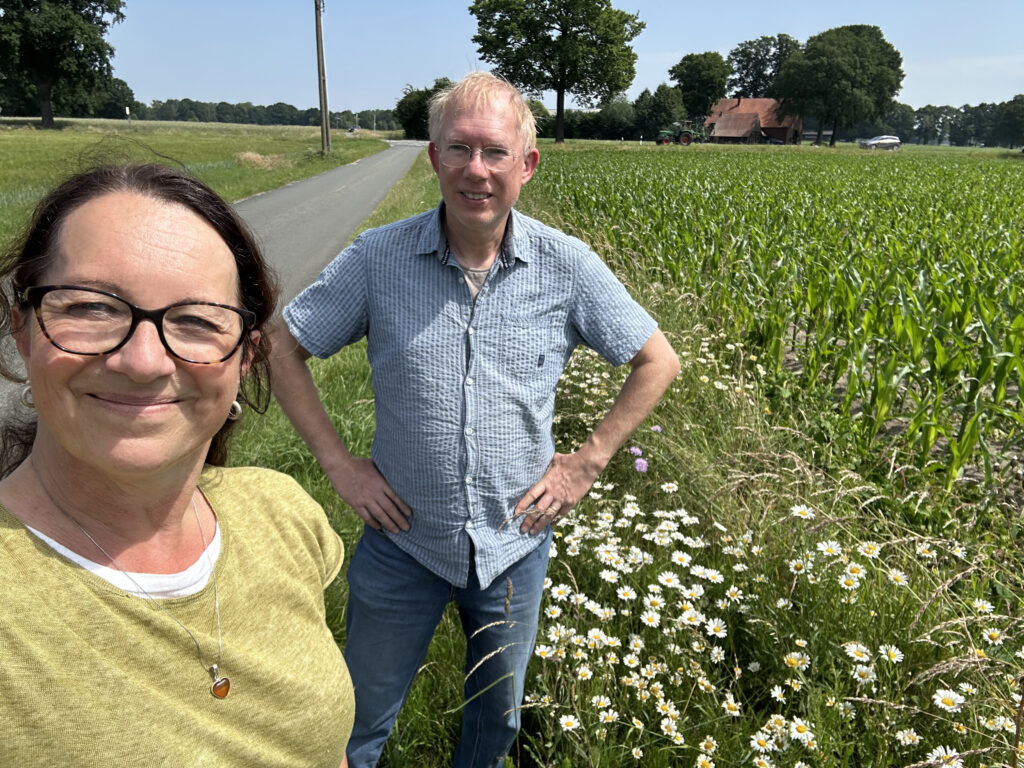 Zwei Personen stehen auf einem Feldweg neben einem Kornfeld und blühenden Gänseblümchen, mit Bäumen und einem Gebäude im Hintergrund bei sonnigem Wetter.