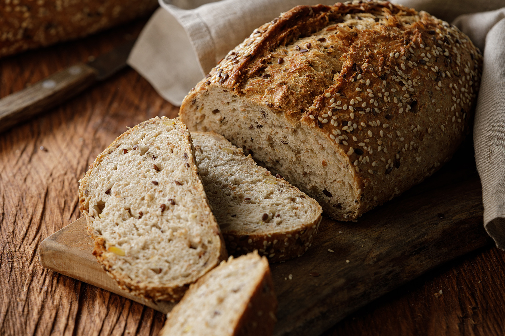 Ein Laib Vollkornbrot, teilweise geschnitten, liegt auf einem Holzbrett, umgeben von einem Leinentuch, mit sichtbaren Samen im Inneren und auf der Kruste.