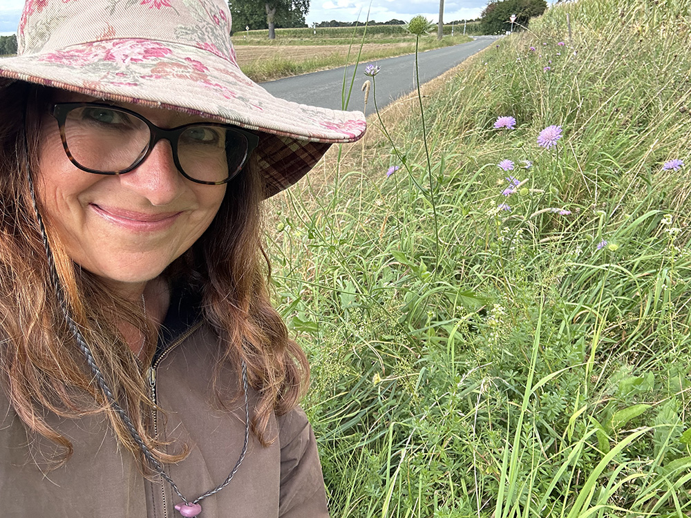 Frau mit Hut und Brille steht lächelnd neben einer wild blühenden Wiese, im Hintergrund eine Landstraße.