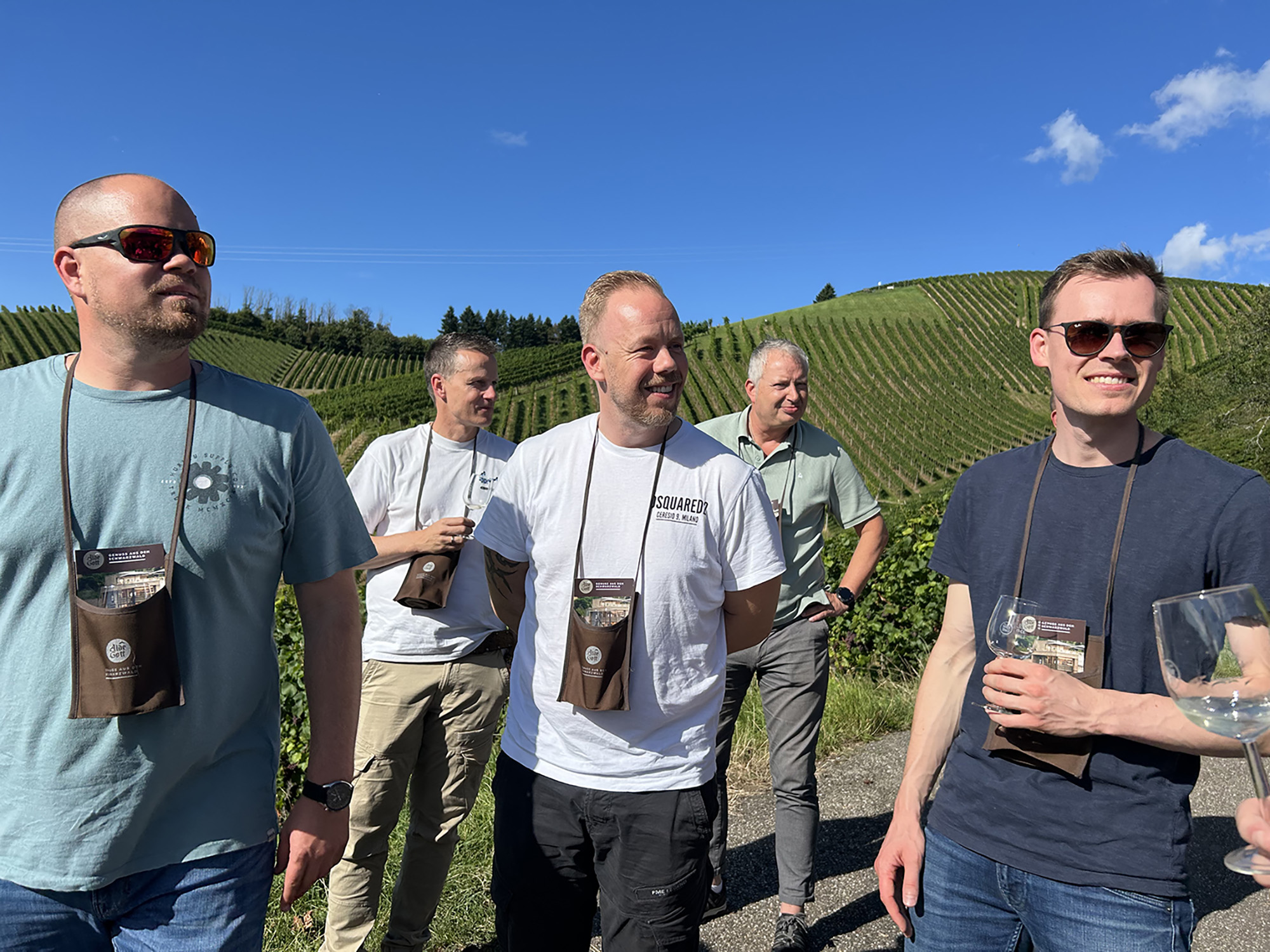 Eine Gruppe von fünf Männern steht unter einem klaren blauen Himmel vor einem Weinberg. Sie tragen lässige Kleidung und Sonnenbrillen und halten Gläser in den Händen. Der Hintergrund zeigt Reihen von Weinstöcken auf einem Hügel.