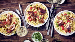 Drei Teller mit Spiralnudeln, gebratener Ente und Tomaten, garniert mit Kräutern, auf einem Holztisch, mit Weingläsern und Besteck.