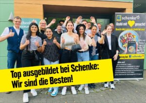 Gruppe von jungen Erwachsenen steht lächelnd vor einem Gebäude, neben einem gelben Schild mit dem Text "Wir sind die Besten!".